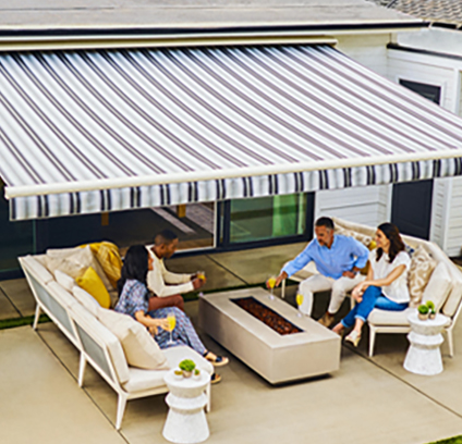 Four people sit on patio furniture around a rectangular fire pit table, beneath a large, striped retractable awning attached to a house, enjoying drinks and conversation on a sunny day.