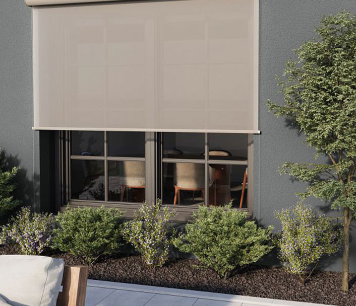 A window with a partially lowered outdoor roller blind, set in a dark gray wall. In front of the window are neatly trimmed shrubs, a small tree, and a tiled patio area. Indoor chairs are visible through the window.
