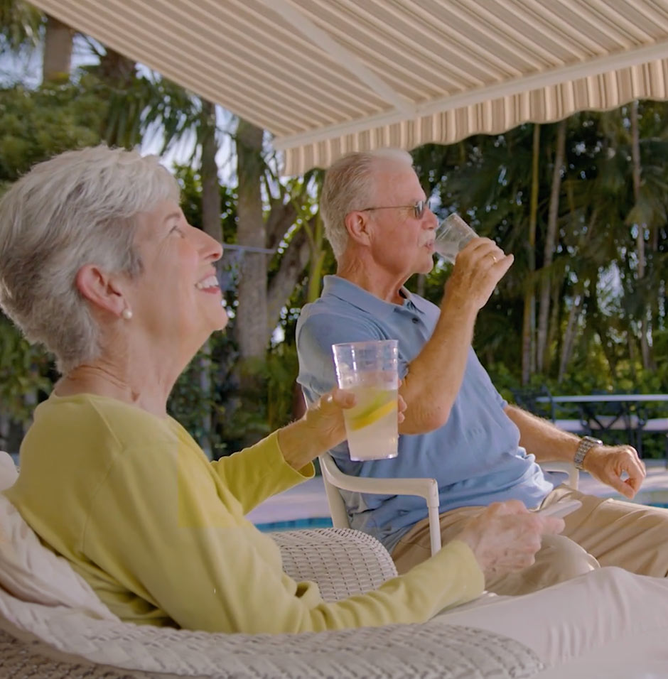 An older woman and man relax outdoors under a striped awning, sitting in wicker chairs by a pool. Both are smiling and holding drinks, with trees and greenery in the background.