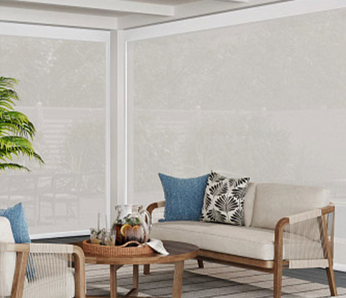 A cozy patio seating area with a sofa and chair featuring wooden frames, beige cushions, blue and patterned throw pillows, a round wooden coffee table with a tray, and a potted plant in the corner.