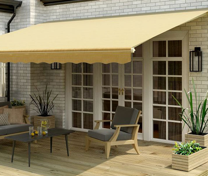 A yellow retractable awning shades a wooden patio with outdoor seating, including a cushioned chair, sofa, a coffee table with drinks, and potted plants near glass double doors.