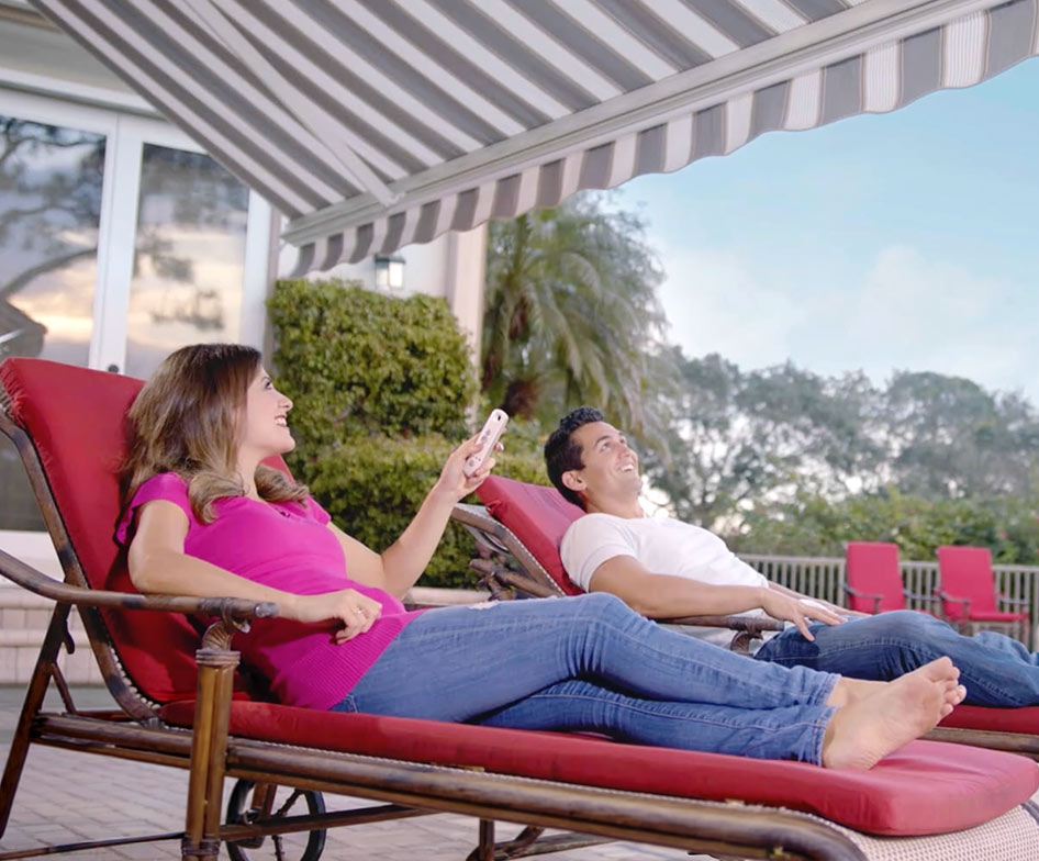 A woman and man relax on red lounge chairs under a striped retractable awning on a patio, with trees and patio furniture in the background. The woman holds a remote control and smiles.