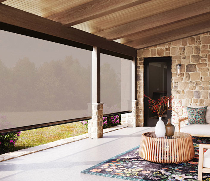 A covered patio with stone walls, large windows with mesh shades, a colorful rug, a round coffee table with vases, and outdoor furniture. Greenery is visible outside through the shades.
