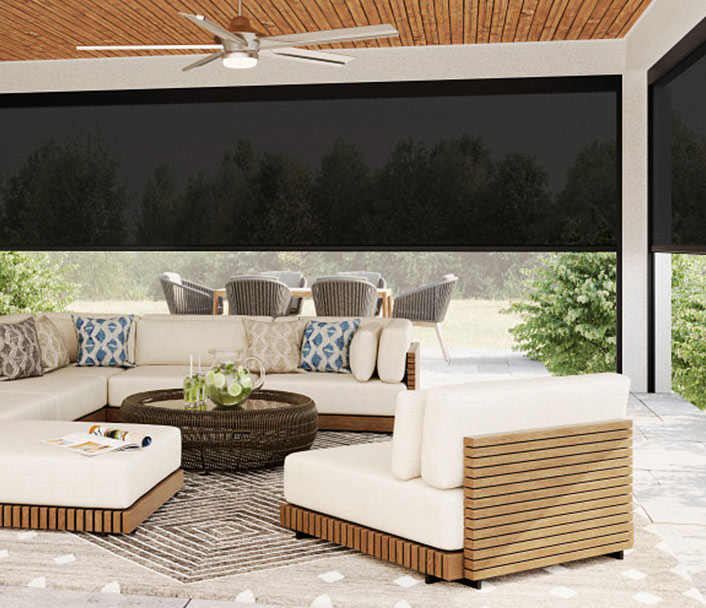 Modern outdoor patio with a large white sectional sofa, patterned pillows, a round wicker coffee table, and a neutral rug. Ceiling fan above and a dining area with gray chairs in the background, surrounded by greenery.