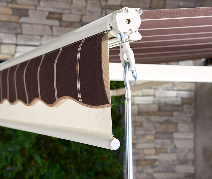 Close-up of a retractable brown and beige striped awning attached to a building with a stone wall and greenery in the background. A metal crank handle is used to open or close the awning.