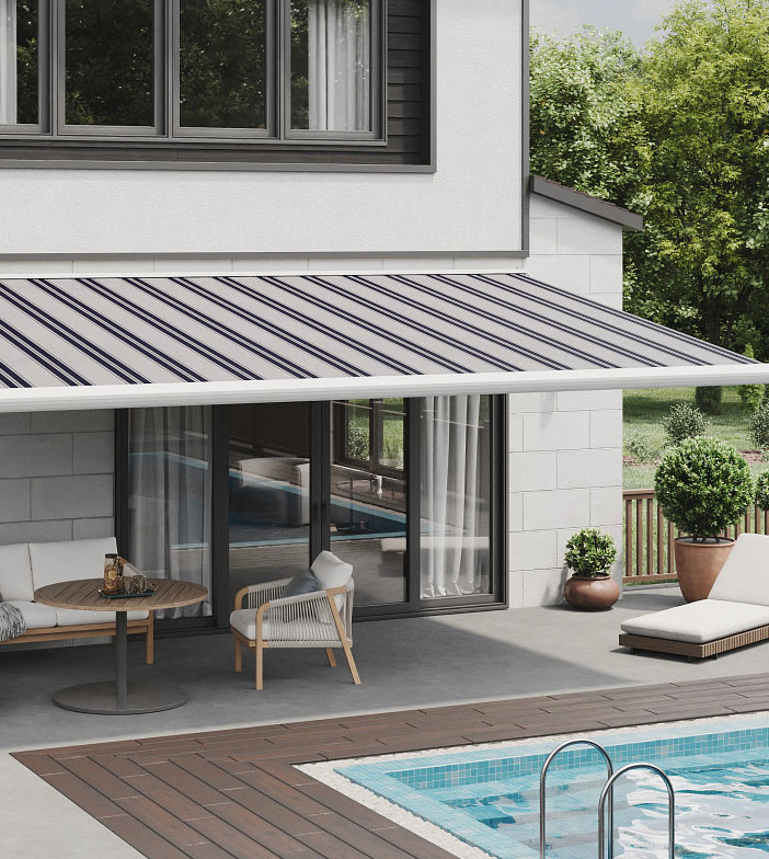 Modern patio with a striped retractable awning, outdoor seating, and a table beside a swimming pool. Glass doors reflect the pool, and potted plants decorate the patio area. Trees are in the background.