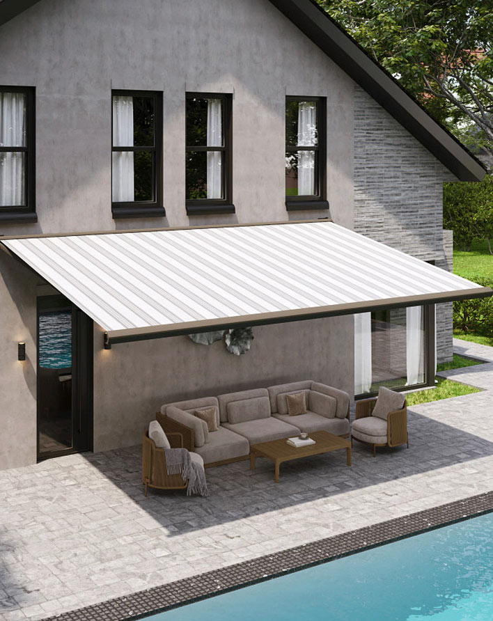 A modern house with a large, striped retractable awning shading a patio area furnished with a beige outdoor sofa set and a wooden coffee table, next to a swimming pool.